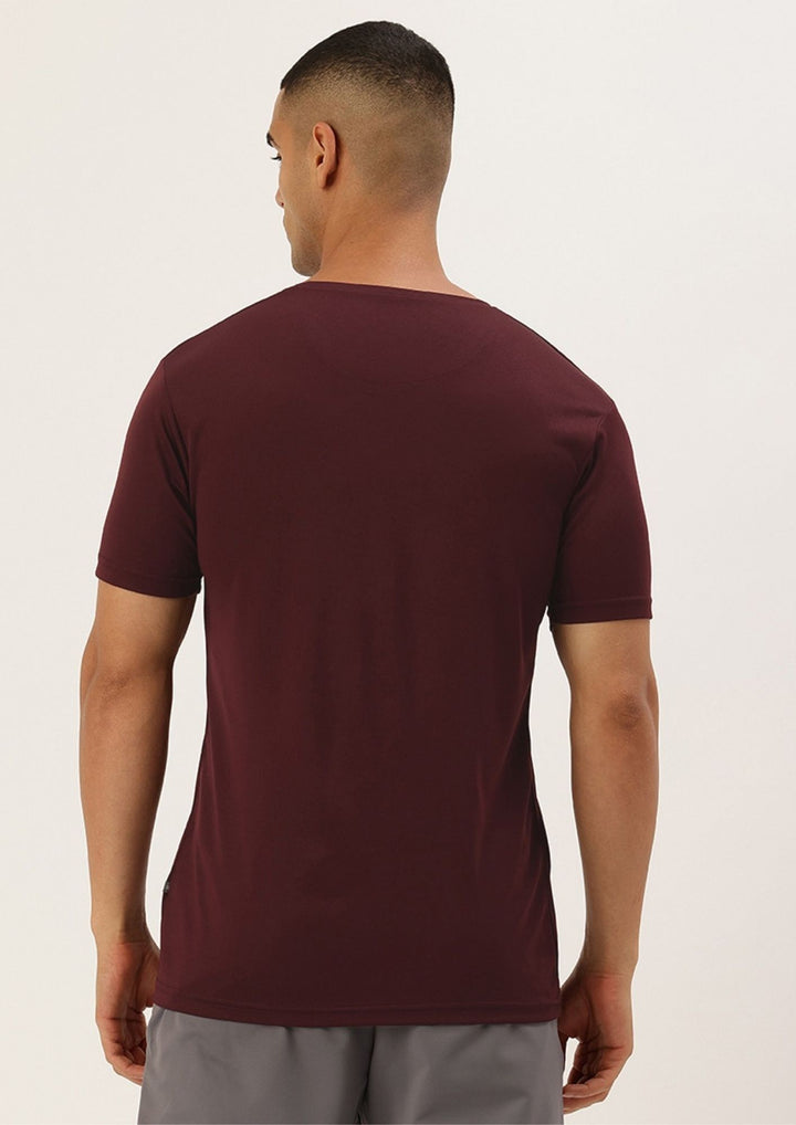 A man viewed from the back wearing a short-sleeve crew-neck t-shirt in maroon, paired with gray pants, against a plain off-white background.