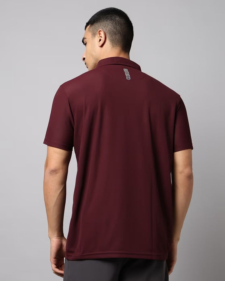 A man viewed from the back wearing a short-sleeve polo shirt in maroon, paired with dark gray trousers, against a solid gray studio background.