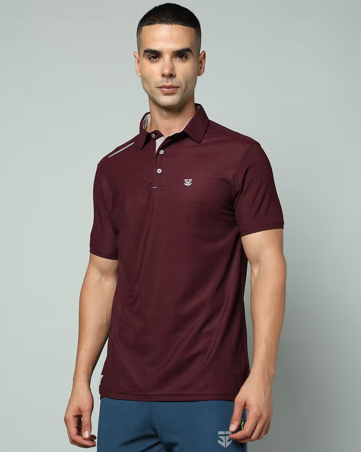 A man wearing a short-sleeve polo shirt in maroon with a small white embroidered logo on the left chest, paired with blue pants, against a plain light gray studio background.