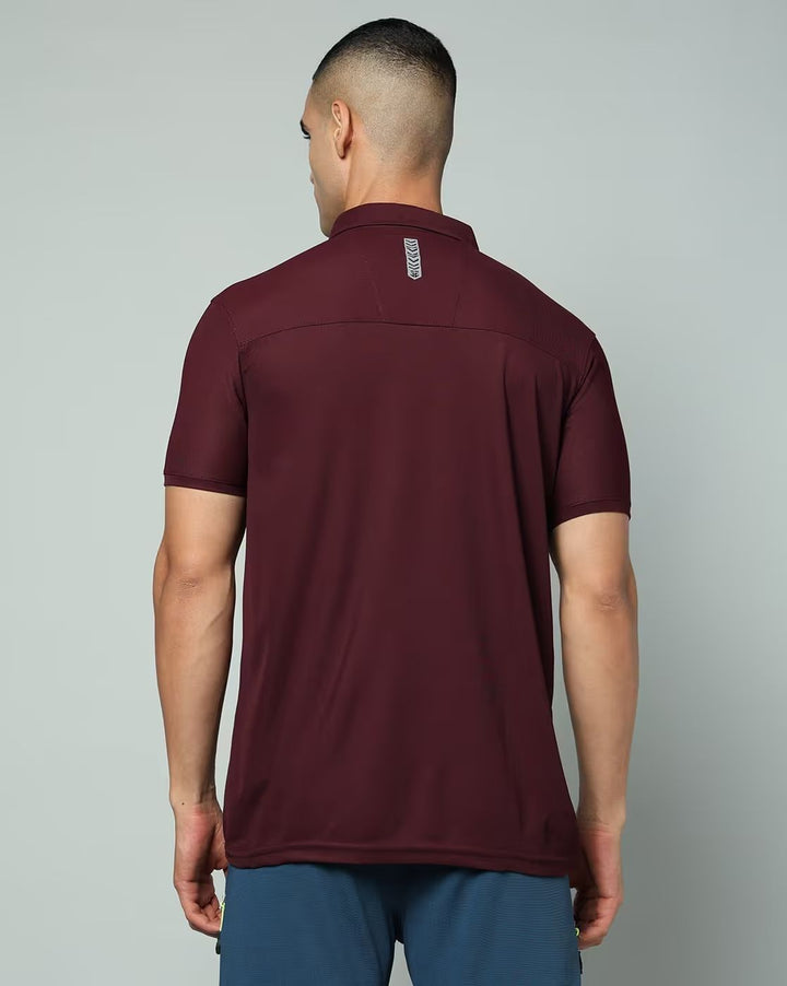 A man viewed from the back wearing a short-sleeve athletic polo shirt in maroon and blue pants, against a plain light gray studio background.