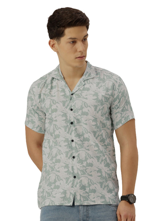 A man wearing a short-sleeve button-up shirt with a light sage green and white tropical floral pattern, paired with blue jeans, against a plain white background.