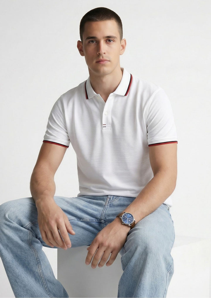 A man wearing a white short-sleeve polo shirt with red trim on the collar and sleeve cuffs, paired with light blue jeans, sitting against a plain light gray background.