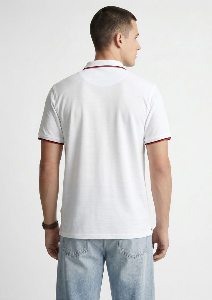 A man viewed from the back wearing a white short-sleeve polo shirt with thin red trim on the collar and sleeve cuffs, paired with light blue jeans against a light gray studio background.