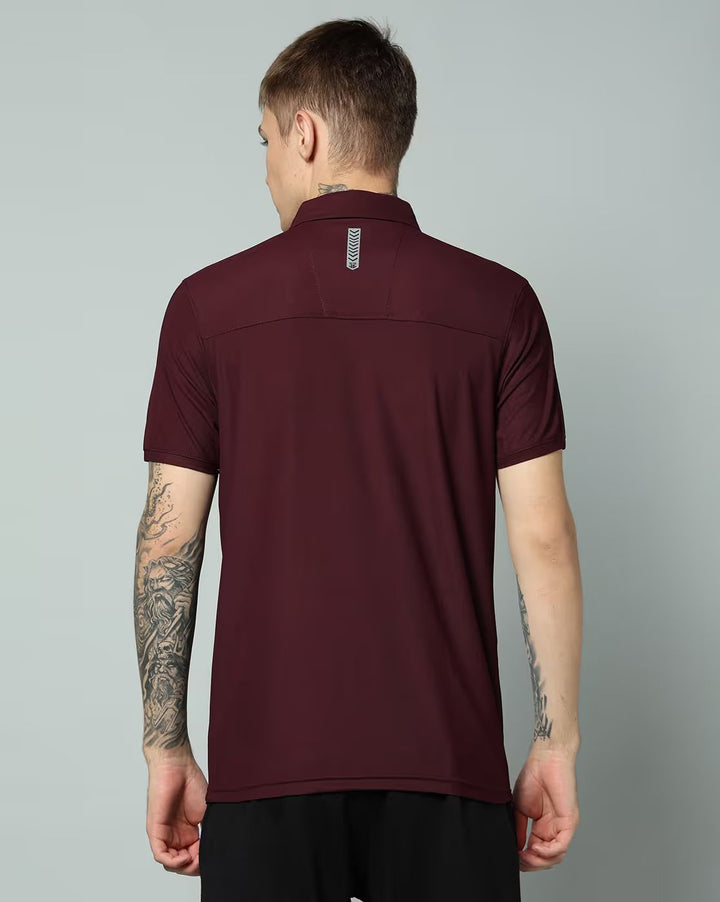 A man viewed from the back wearing a short-sleeve athletic polo shirt in maroon with a small vertical reflective logo patch at the collar, paired with black shorts, against a solid light gray studio background.