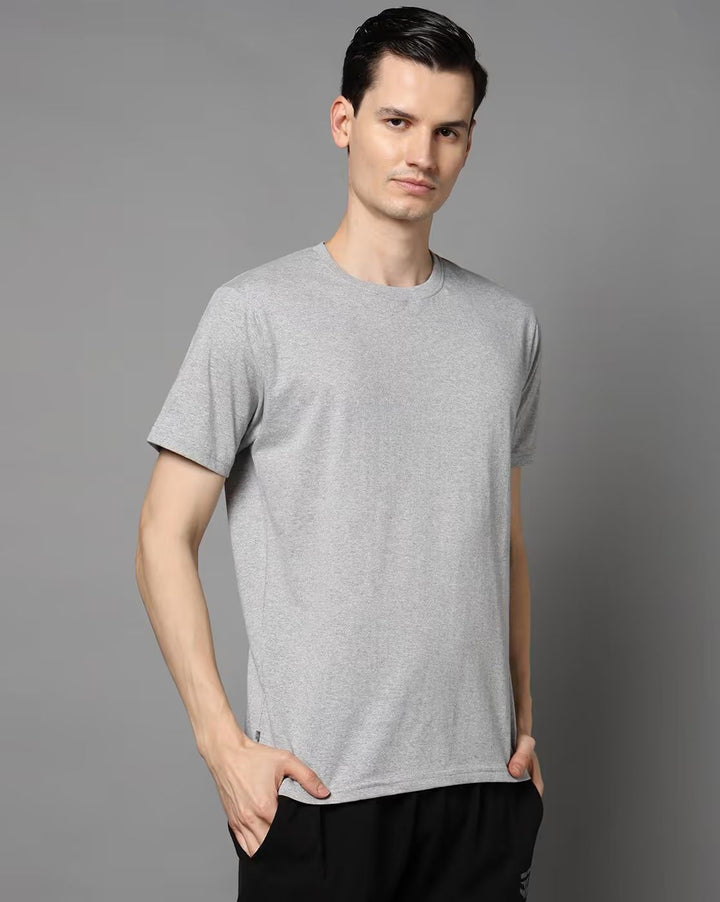 A man wearing a light gray short-sleeve crewneck t-shirt and black pants, standing against a solid gray studio background.