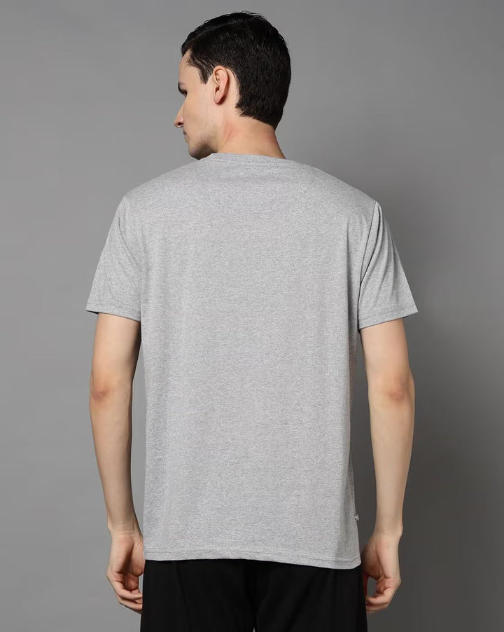 A man viewed from the back wearing a light gray short-sleeve crewneck t-shirt and black pants, standing against a solid gray studio background.