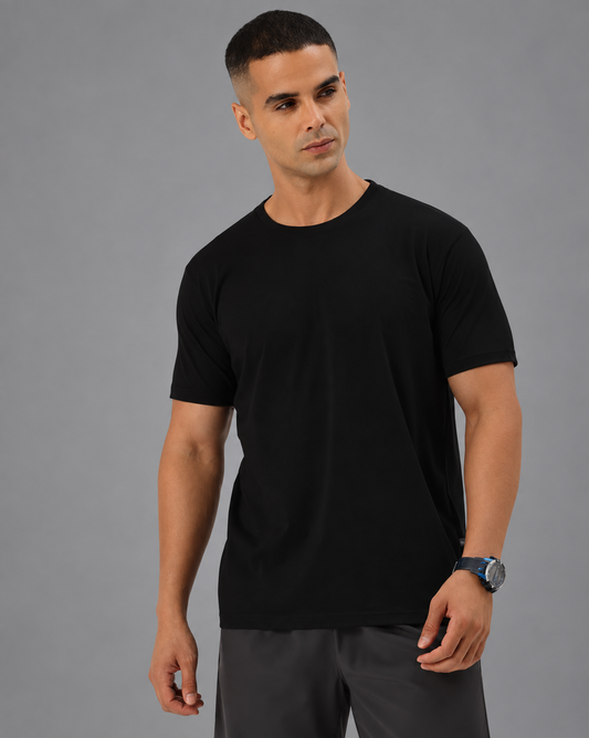 A man wearing a black short-sleeve crewneck t-shirt and grey pants, standing against a solid gray studio background.