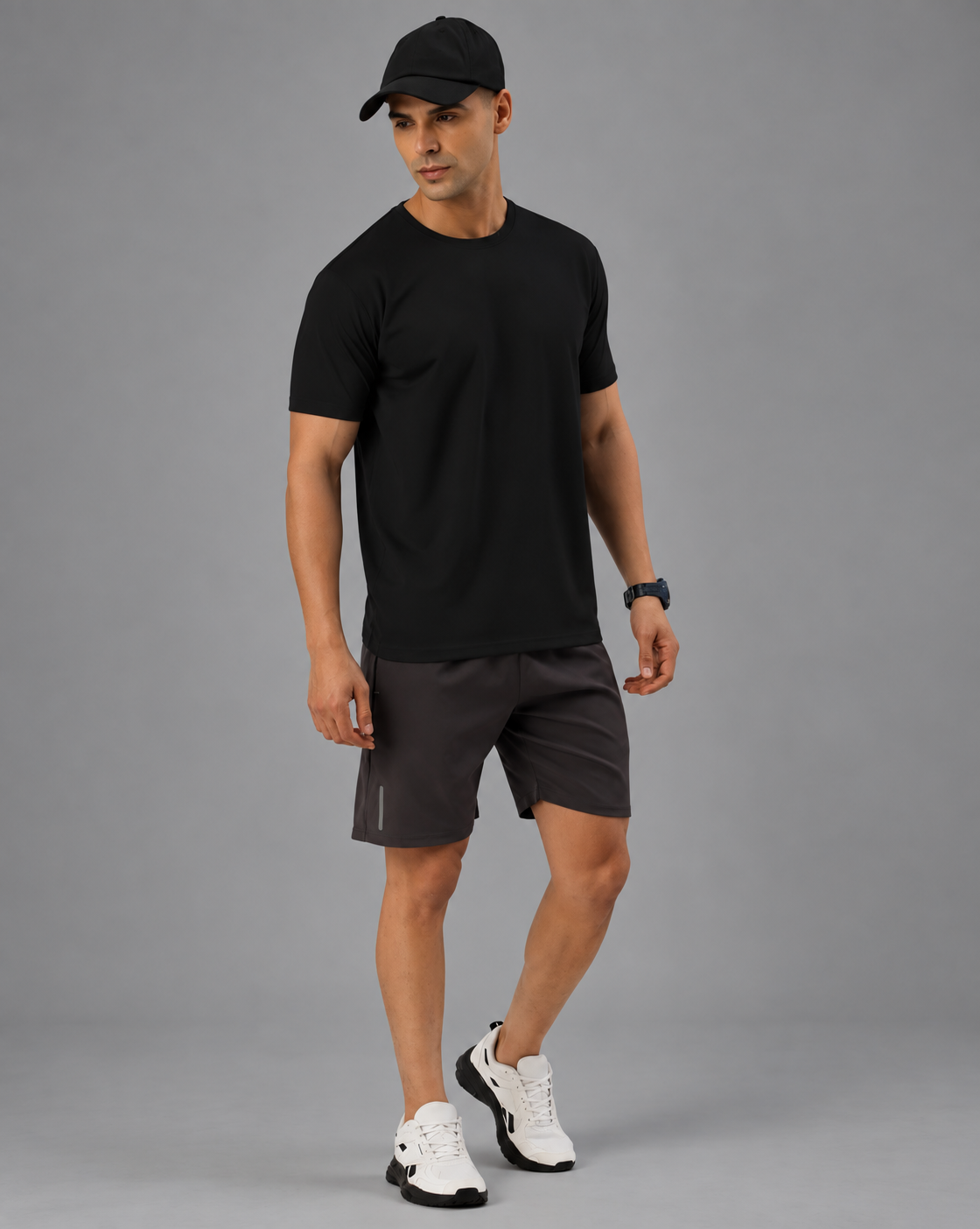 A man wearing a black short-sleeve crewneck t-shirt and grey shorts , standing against a solid gray studio background.