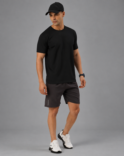 A man wearing a black short-sleeve crewneck t-shirt and grey shorts , standing against a solid gray studio background.