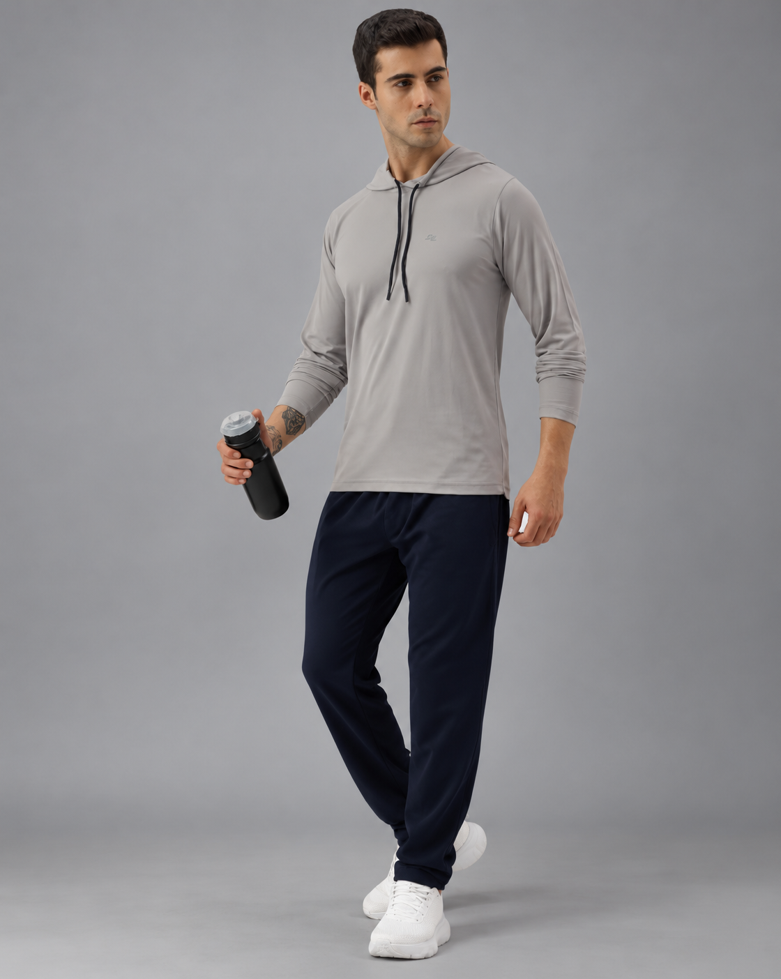 A man modeling a light gray long-sleeve athletic hoodie with dark navy jogger pants and white sneakers, holding a black water bottle against a plain greybackground.