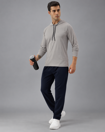 A man modeling a light gray long-sleeve athletic hoodie with dark navy jogger pants and white sneakers, holding a black water bottle against a plain greybackground.
