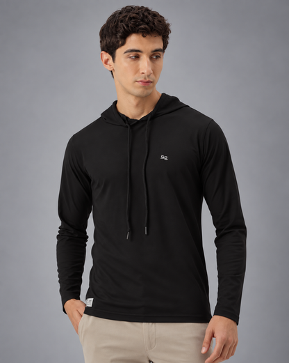 A man wearing a black long-sleeve athletic hoodie with drawstrings and a small white logo on the left chest, paired with light beige pants, against a plain grey background.