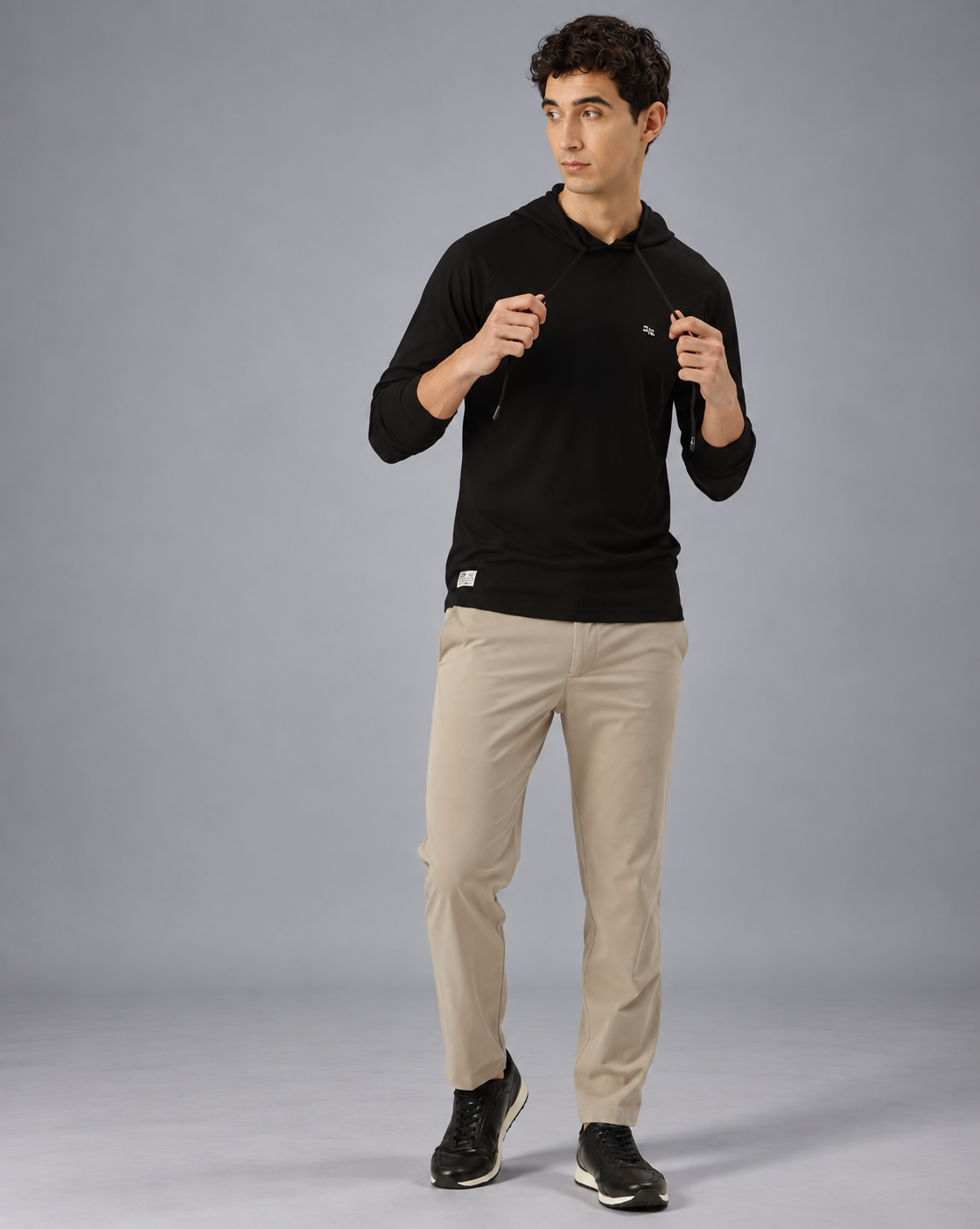 A man wearing a black long-sleeve athletic hoodie with drawstrings and a small white logo on the left chest, paired with light beige pants, against a plain grey background.