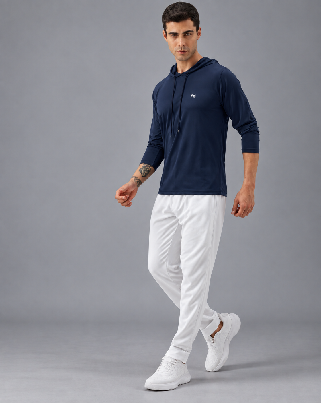 A man modeling a dark navy long-sleeve hoodie with a small white embroidered logo on the left chest, paired with white joggers and white sneakers, set against a plain white background.