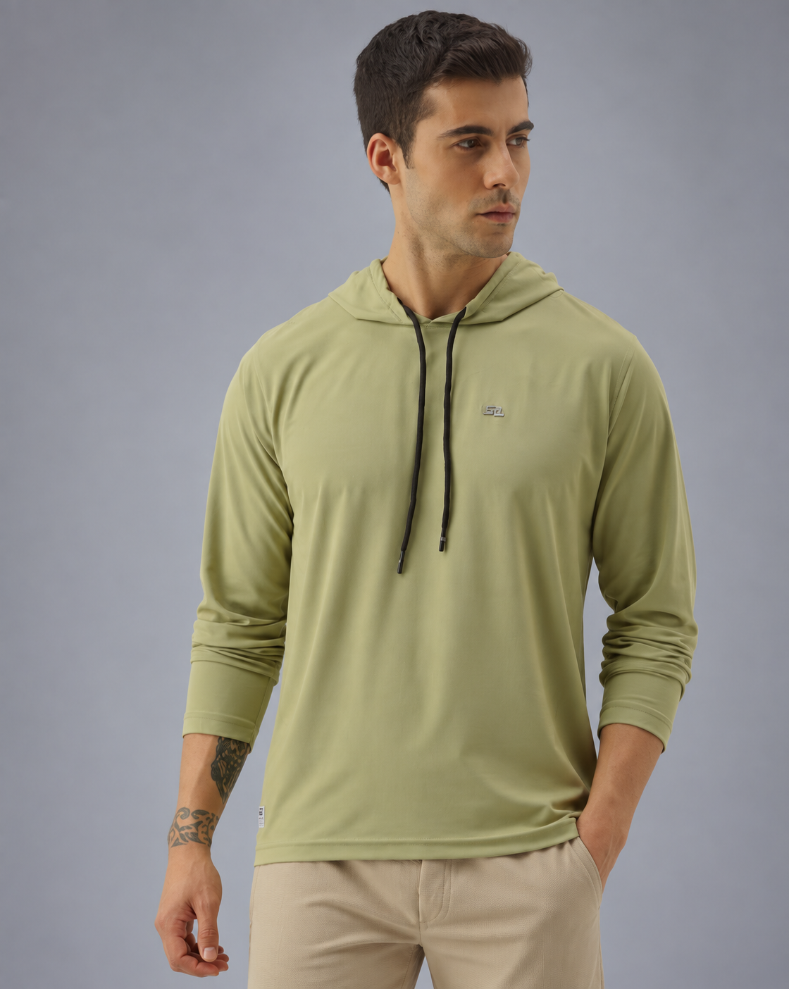 A man wearing a sage green long-sleeve hooded athletic shirt with black drawstrings and beige pants, standing against a solid gray studio background.
