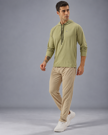 A man modeling a sage green long-sleeve athletic hoodie with black drawstrings and beige jogger pants, paired with white sneakers, against a solid gray studio background.